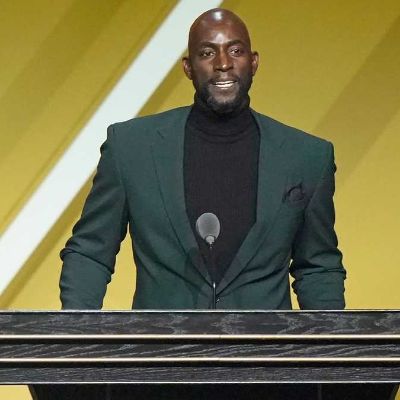 He is standing in front of the podium wearing  a black suit and a turtleneck.